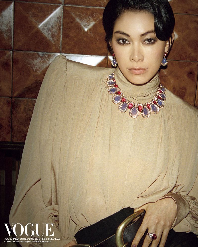 Elegant Asian woman in neutral blouse, statement jewelry, and black skirt, posed against a textured brown backdrop in a studio setting.
