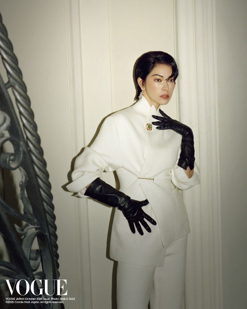 Elegant Asian woman in a white pant suit and black gloves poses indoors near a staircase for a fashion editorial style photo.