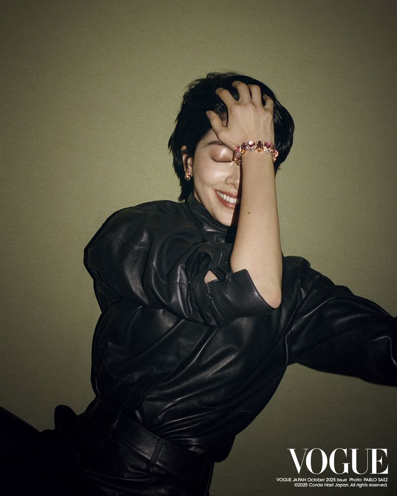 Smiling woman with short dark hair and a black leather jacket poses with her hand on her head in a studio shot.