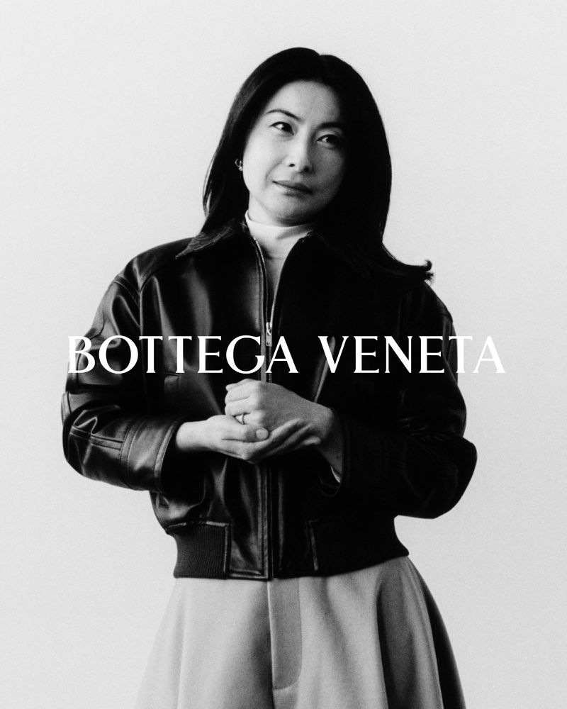 Black-and-white portrait of an Asian woman wearing a black leather jacket and light-colored trousers, posing against a white background.