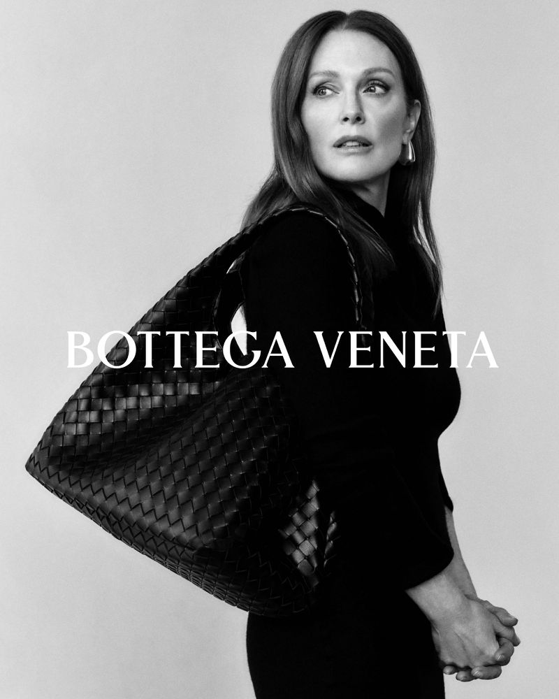 Elegant portrait of an adult woman with long brown hair, wearing a black dress, posing with a woven leather handbag in a studio.