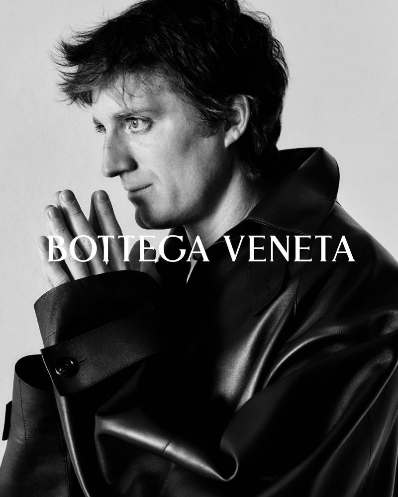 Black and white portrait of a man wearing a dark coat. He gazes off to the side with his hands clasped under his chin, Bottega Veneta brand text.