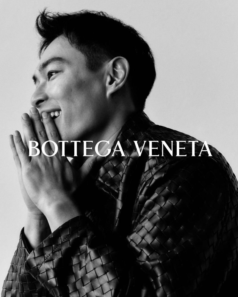 Close-up black and white portrait of a smiling Asian man in a woven jacket, with his hands near his face and the Bottega Veneta logo.