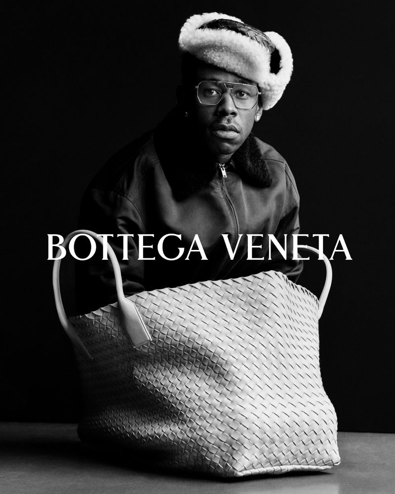 Tyler, The Creator posing indoors wearing fur hat, jacket, glasses next to woven Bottega Veneta handbag on table in black and white.