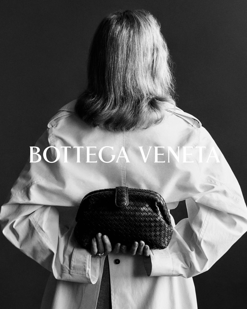 Woman poses from the back wearing trench coat, holding Bottega Veneta woven clutch. Monochrome fashion advertisement.