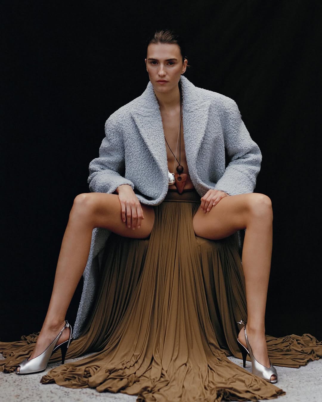 A woman sits in a draped skirt, wearing a coat and heels against a dark backdrop, showcasing a fashion editorial style.