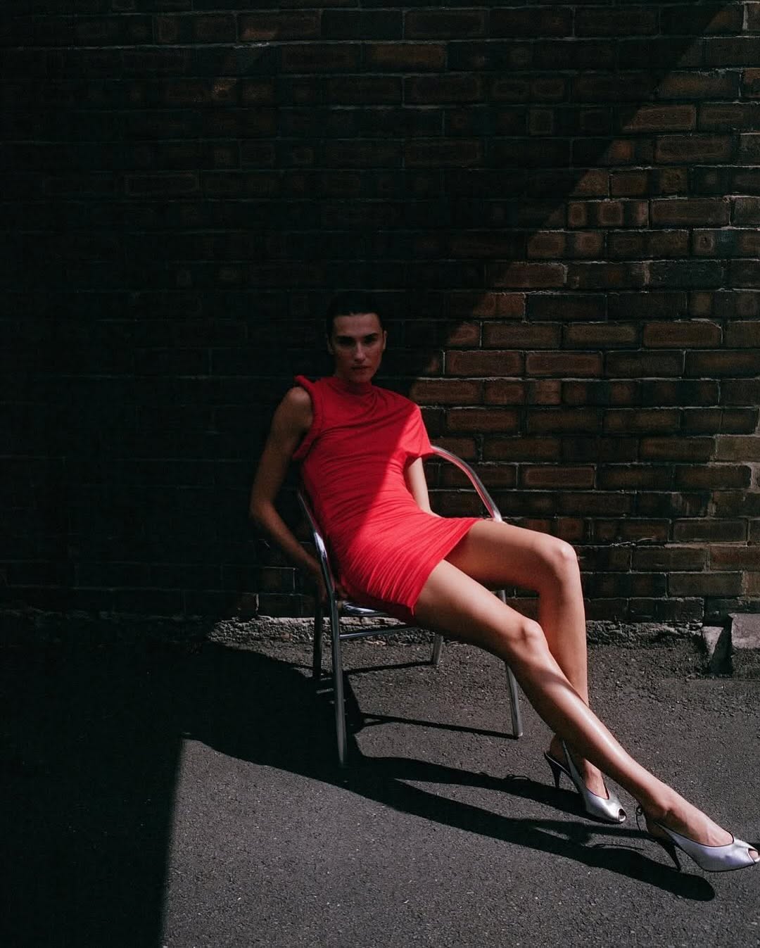 A woman poses in a red dress and heels, seated on a chair against a brick wall with dramatic lighting and shadows creating a fashion editorial look.