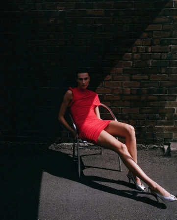 A woman poses in a red dress and heels, seated on a chair against a brick wall with dramatic lighting and shadows creating a fashion editorial look.