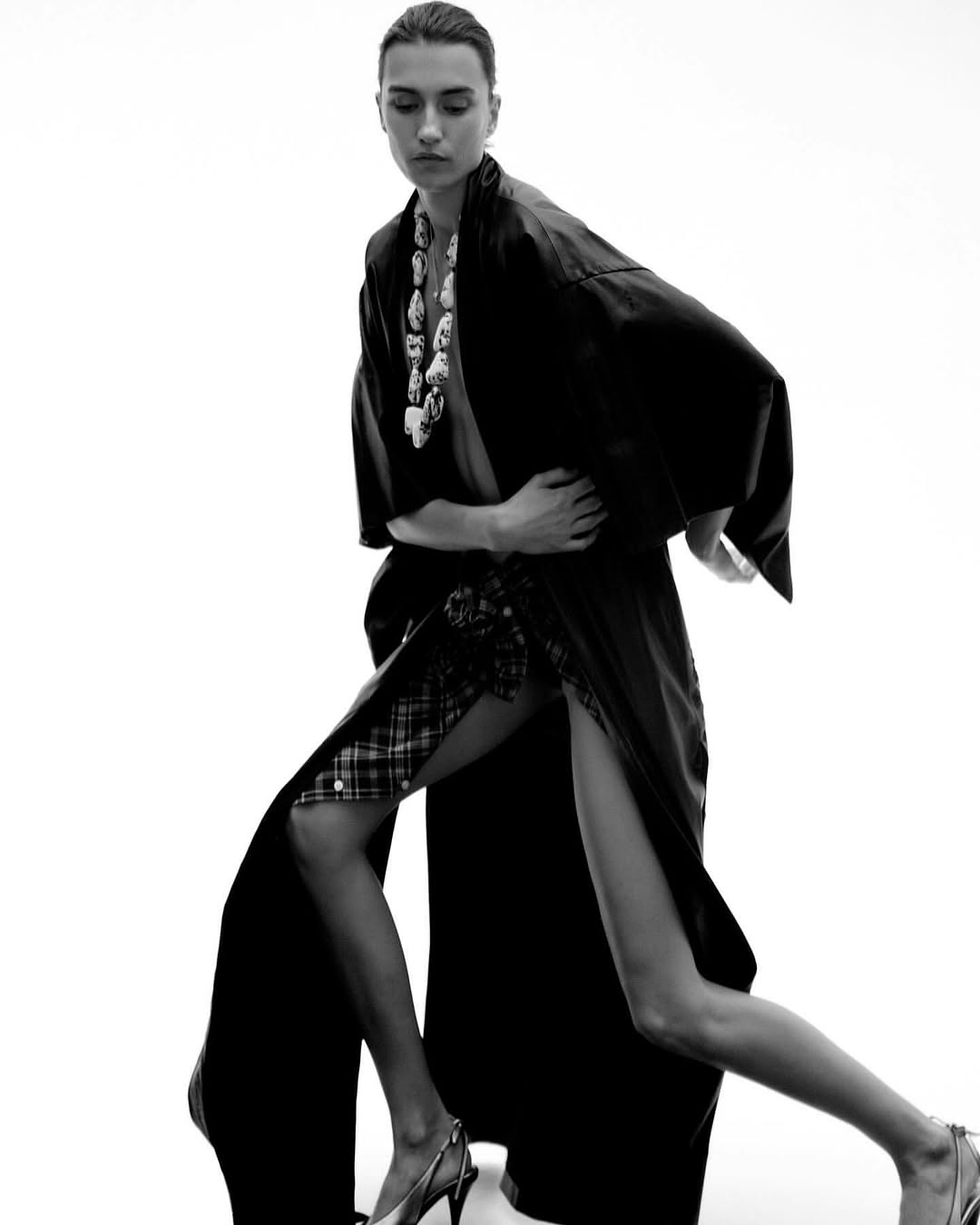 Monochrome fashion shot of a model in a black robe with a high slit, necklace, and heels posing against a white background.