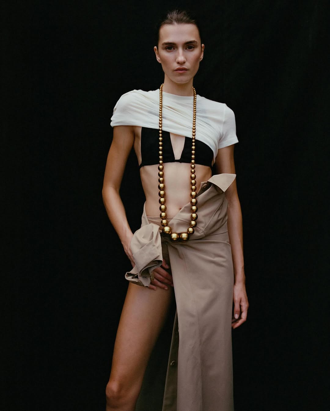 Fashion portrait of a woman with tan trench coat unbuttoned, large gold bead necklace, white crop top, and black bra against a black backdrop.
