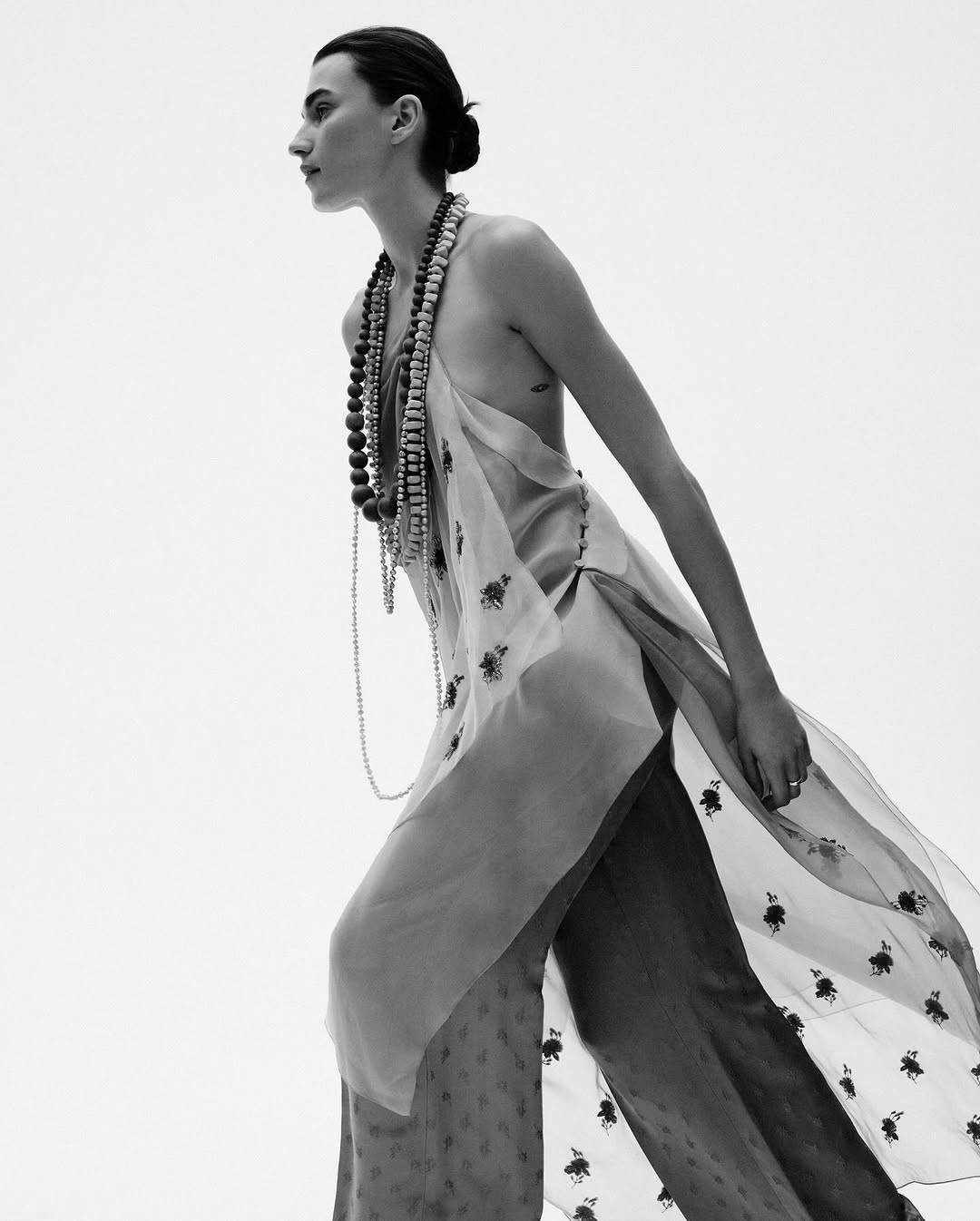 A woman in a long, sheer top and wide-leg pants stands against a white background, adorned with strands of beads in a fashion-forward editorial shot.