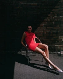 Model wearing a bright red dress and silver heels seated on a metal chair against a dark brick wall exterior.