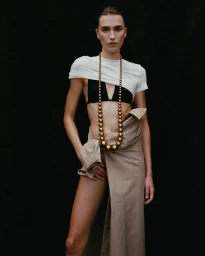 Model in white top and beige skirt wearing a long gold bead necklace against a dark background for COAT Magazine