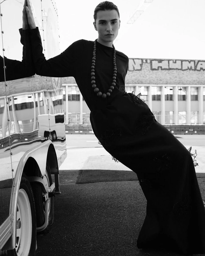 Model leaning against a reflective vehicle exterior wearing a dark long-sleeve top, maxi skirt, and beaded necklace