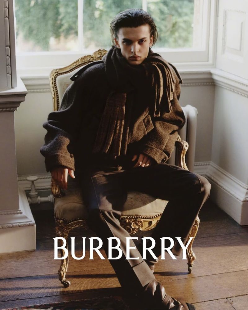 A fashion shot featuring a young adult male model with brown hair in a coat and scarf seated on an ornate chair in a room with hardwood floors.