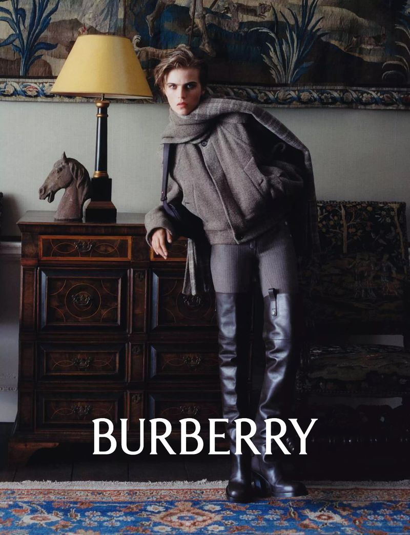 Fashionable adult woman in Burberry outerwear and thigh-high leather boots standing in an elegant interior setting near a lamp.