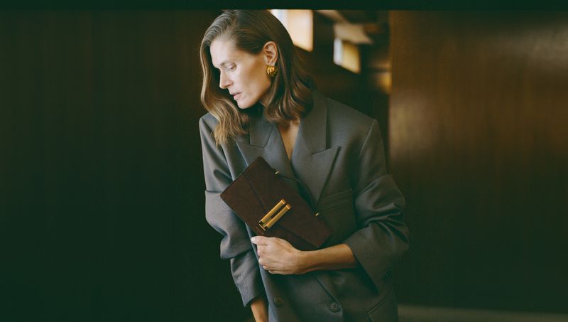 Adult woman in a grey blazer holding a brown clutch bag, posed indoors. Fashion editorial shot with neutral tones and soft lighting.