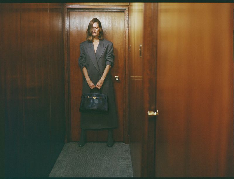 Malgosia Bela poses in a wood-paneled hallway holding a black leather tote handbag while wearing an oversized charcoal grey coat.