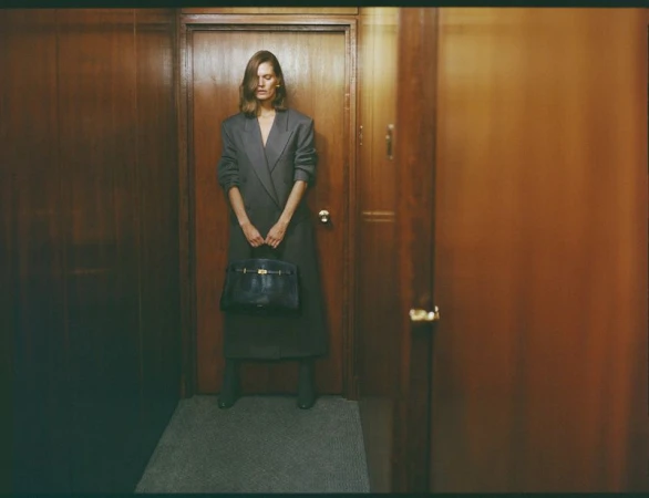 Malgosia Bela poses in a wood-paneled hallway holding a black leather tote handbag while wearing an oversized charcoal grey coat.