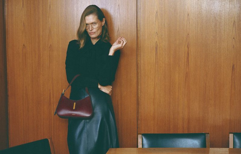 Adult female poses in front of wood panel wall with handbag wearing skirt, sweater, and eyeglasses in an eye-level medium shot.
