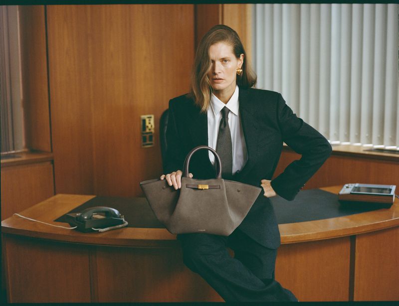 A stylish adult woman poses in a business suit with a structured leather handbag in an indoor office setting, exhibiting fashion and professionalism.