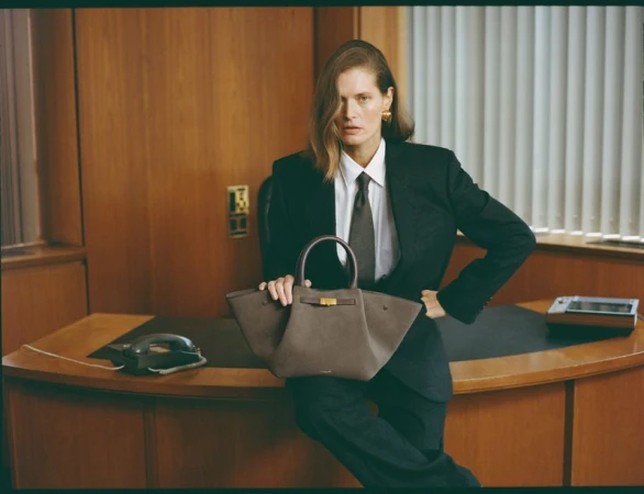 A stylish adult woman poses in a business suit with a structured leather handbag in an indoor office setting, exhibiting fashion and professionalism.