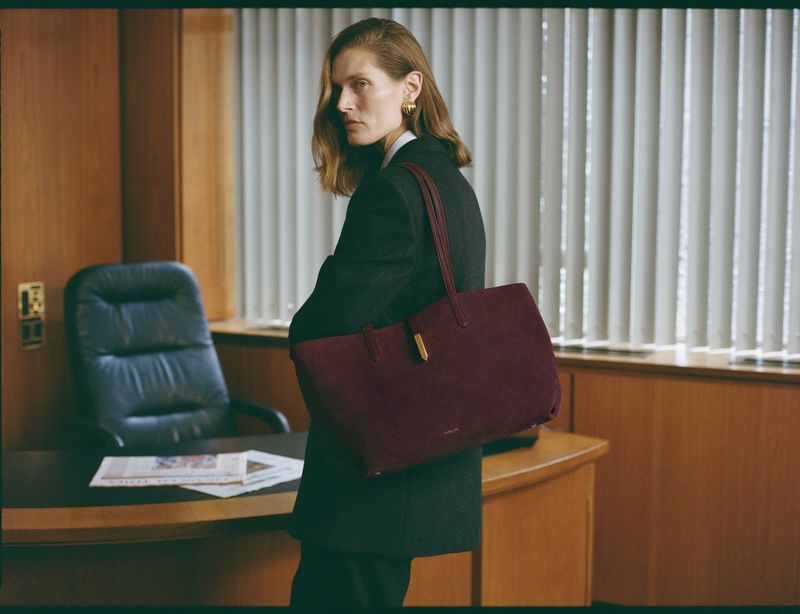 Adult female model poses indoors wearing a black blazer and carrying a burgundy tote bag, with a desk and office chair visible.