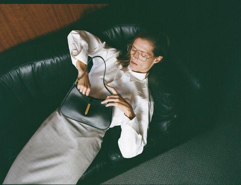 Fashionable woman on a black leather sofa with a black purse, wearing a long grey skirt and white blouse, stylish glasses, indoor lighting.