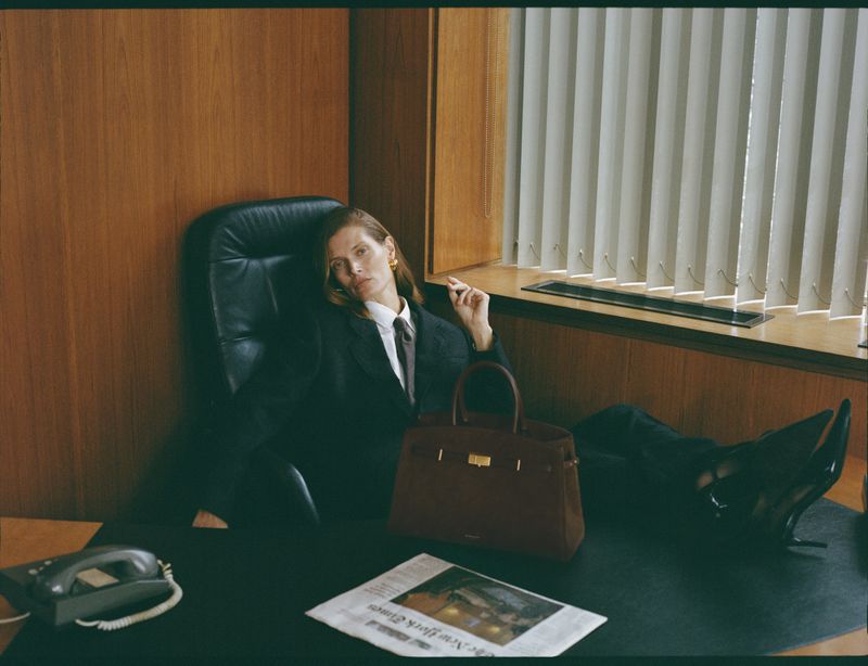 Woman in black suit reclines on desk with handbag and newspaper in office setting. She is wearing gold earrings and a tie.