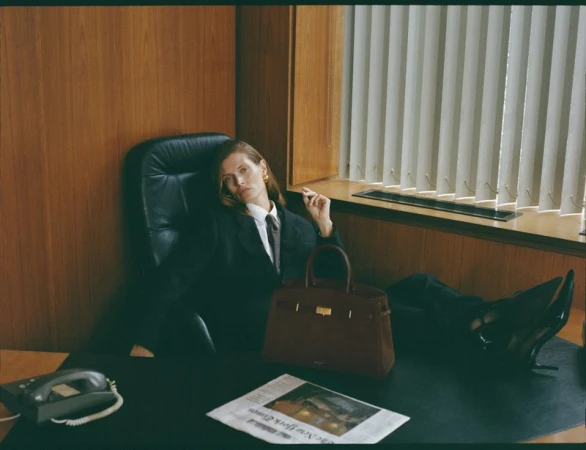 Woman in black suit reclines on desk with handbag and newspaper in office setting. She is wearing gold earrings and a tie.