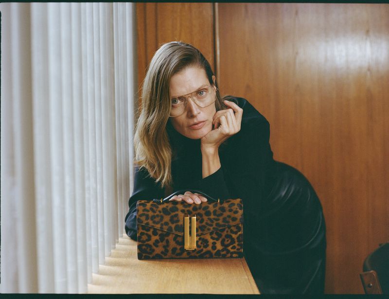 Elegant blonde woman poses indoors with a leopard print handbag, wearing glasses and a black turtleneck, seated against wood paneling.