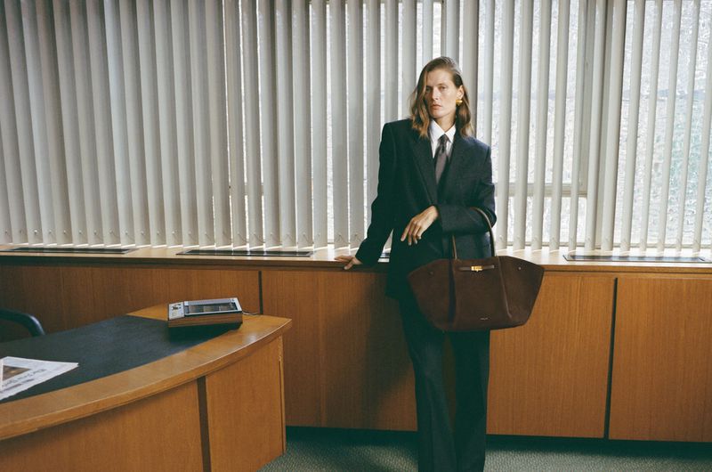 Woman in a dark suit holding a brown leather tote bag, standing in a vintage office setting with blinds and a wood desk.