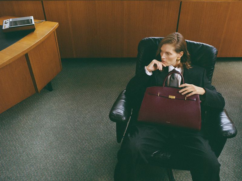 Woman in business attire sits in an office chair holding a burgundy leather handbag, with an office desk visible in the background.