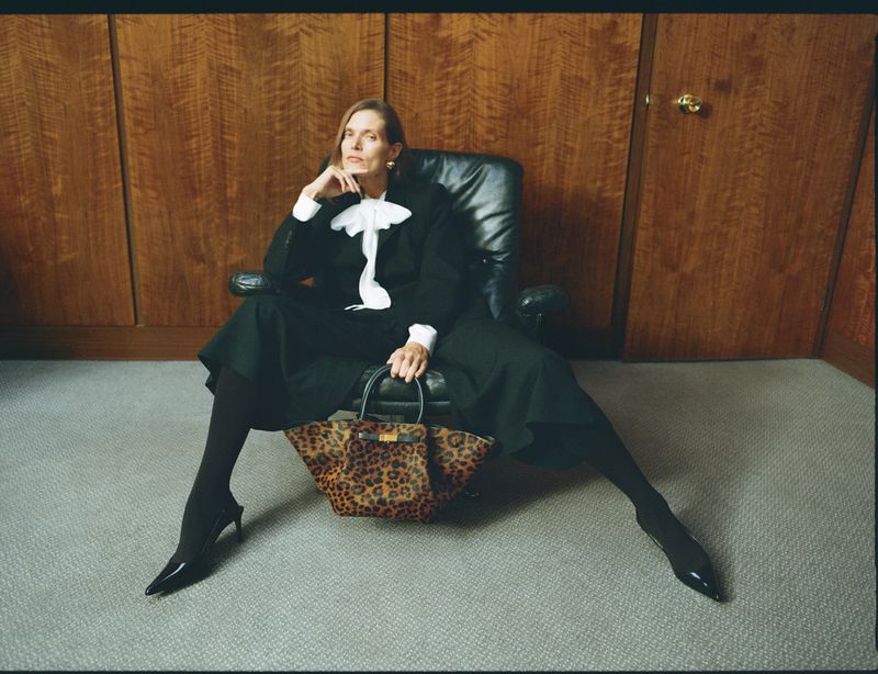 Elegant older woman in a black suit sits in a leather chair with a leopard-print handbag, posing against a wood-paneled wall, style and sophistication.