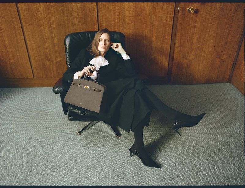 Elegant woman in a black blazer and skirt sits in a leather chair, holding a brown handbag in a wood-paneled room with gray carpet.