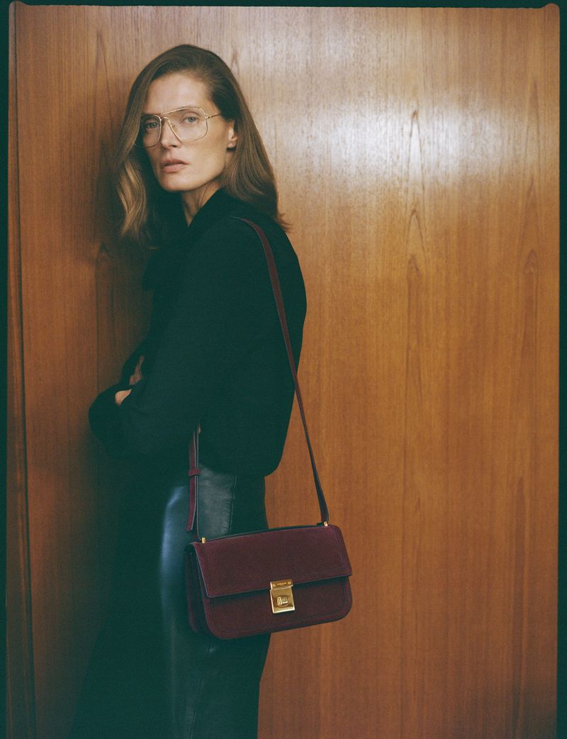 Portrait of a woman with glasses wearing a black top and carrying a burgundy purse in front of a wooden wall.