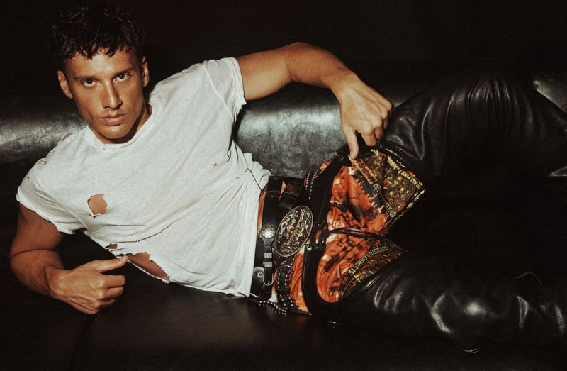 Stylish man in ripped t-shirt and leather pants poses on black sofa in dim light, showcasing unique belt and confident pose.