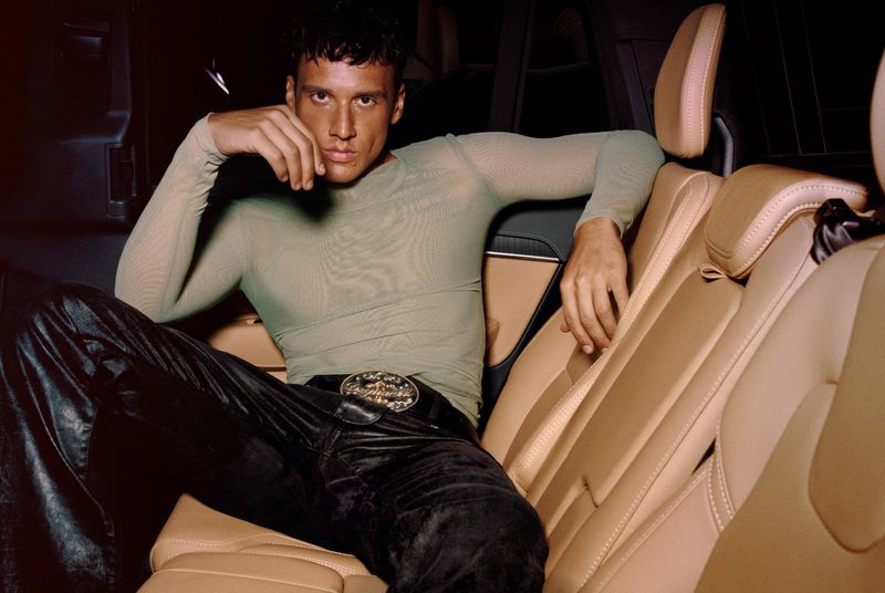 Adult male poses in a car's back seat with tan leather interior and a large decorative belt buckle, wearing a sheer top and dark pants.