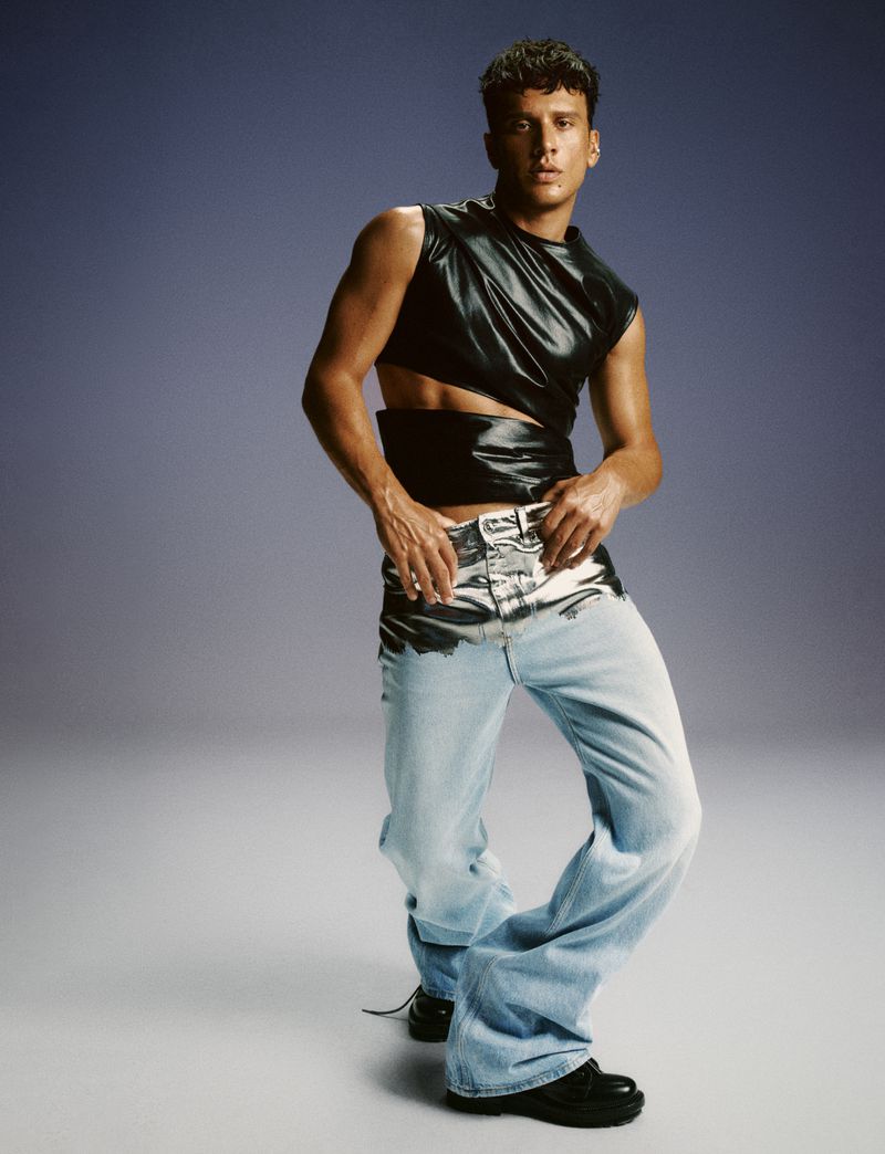 Man poses in studio wearing a black sleeveless cutout top and light blue jeans with silver accents, set against a grey gradient backdrop.