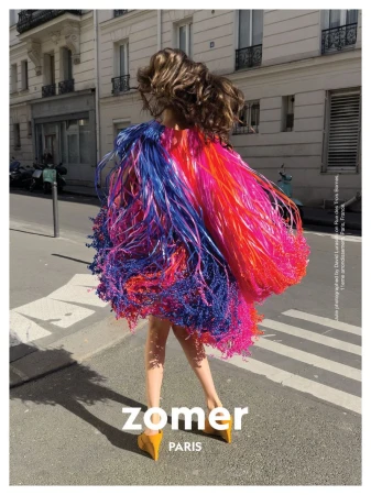 A fashion photo showcases a person in a vibrant, fringed garment, standing on a city street with buildings in the background on a sunny day.