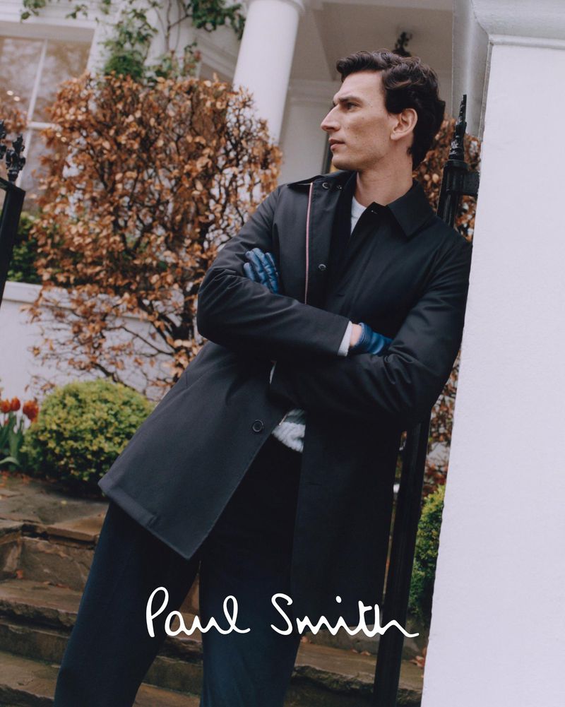 A dark-haired man models a dark navy trench coat with patterned blue gloves, outside a white building, for a Paul Smith fashion campaign.
