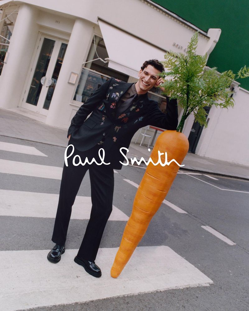 A smiling adult male model wearing a patterned suit leans against a giant carrot on a crosswalk outdoors, with the Paul Smith logo.