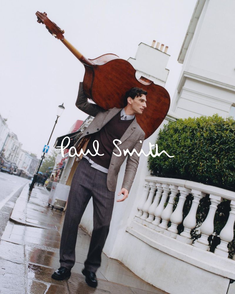 Fashionable man carrying an upright bass on a rainy street with white building and 'Paul Smith' logo overlay, outdoor shot.