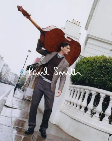 Fashionable man carrying an upright bass on a rainy street with white building and 'Paul Smith' logo overlay, outdoor shot.