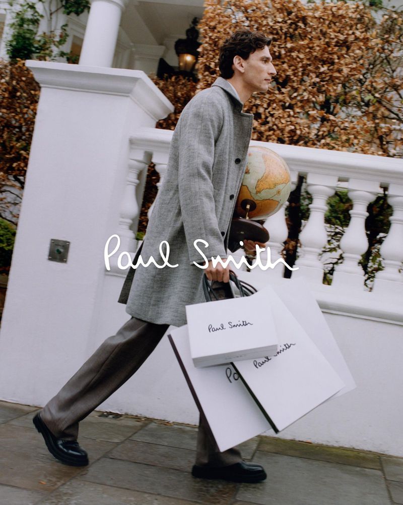 A man in a grey coat walks on a sidewalk carrying shopping bags and a globe in a promotional outdoor fashion shot for Paul Smith.