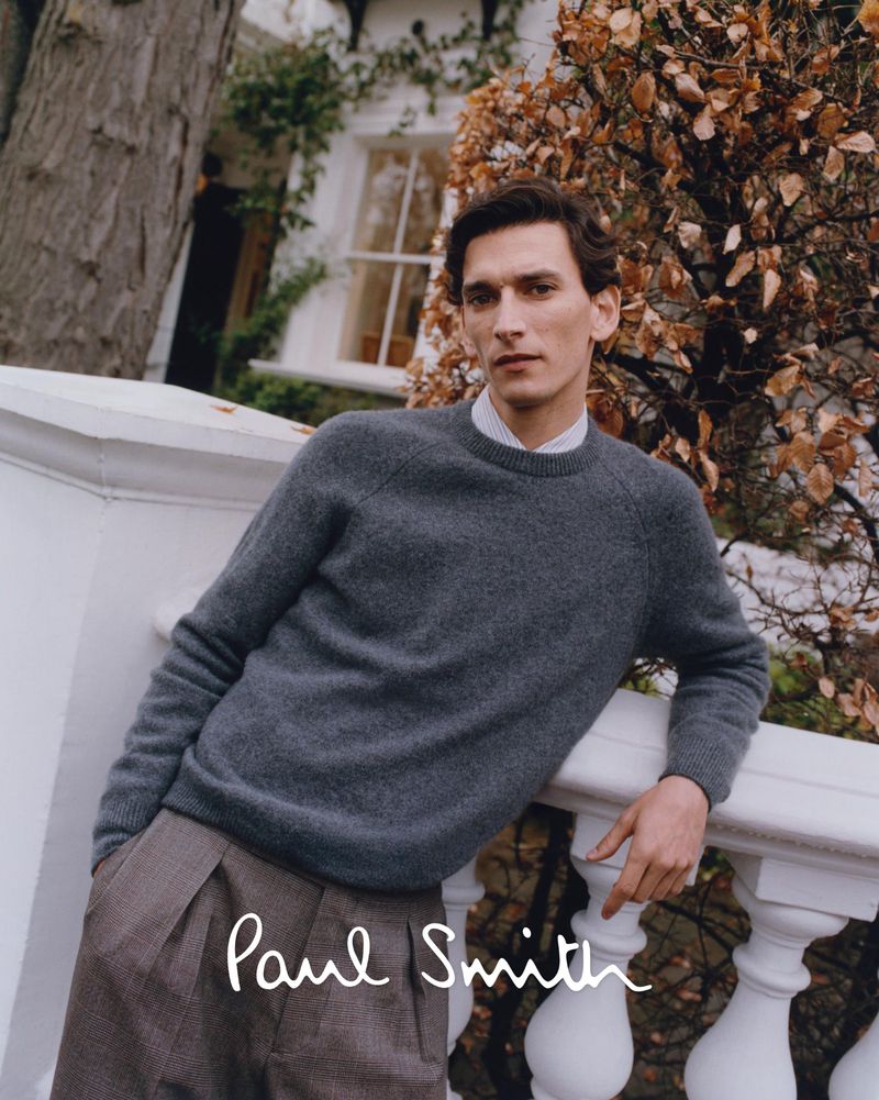 A handsome man poses outdoors in a grey sweater and brown pleated pants, leaning on a white balustrade with a tree in the background.