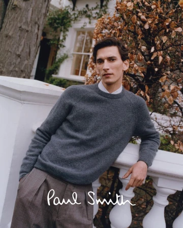 A handsome man poses outdoors in a grey sweater and brown pleated pants, leaning on a white balustrade with a tree in the background.