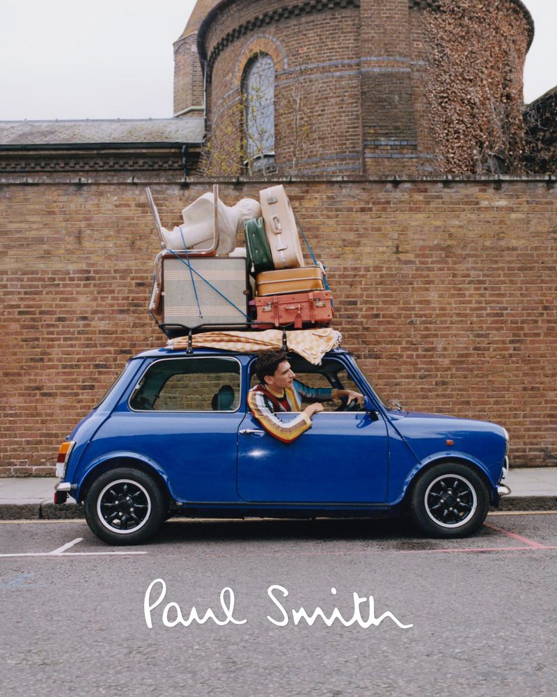 Blue Mini Cooper with luggage on top driving down a street in front of a brick wall, next to a brick building under an overcast sky.