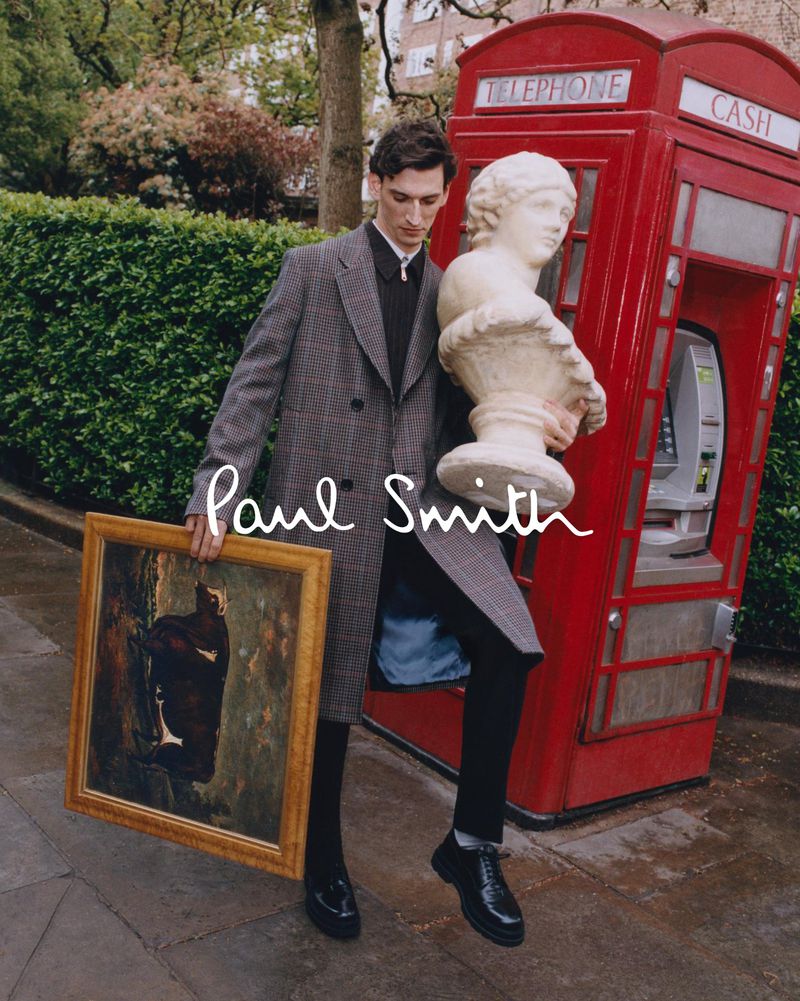 Male fashion model carries art and sculpture in outdoor editorial setting near London telephone booth with muted, desaturated color grade.