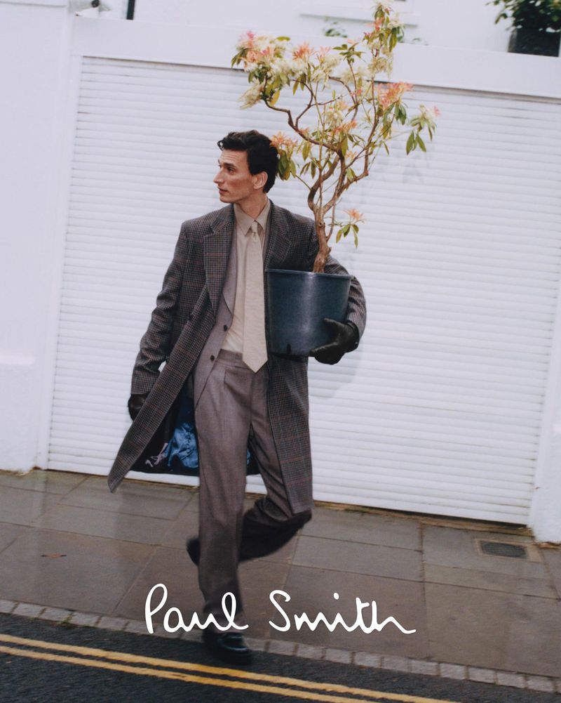 A man in a grey suit and coat carries a potted plant in front of a white garage door, showcasing a blend of fashion and nature.